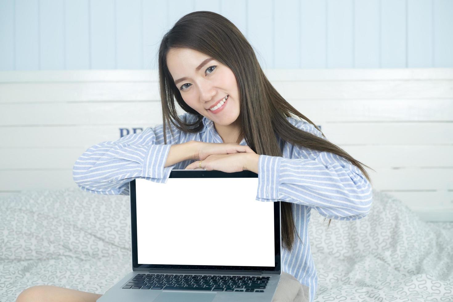 Joven mujer asiática sonriendo y mostrando la pantalla de la computadora portátil en blanco en su dormitorio foto