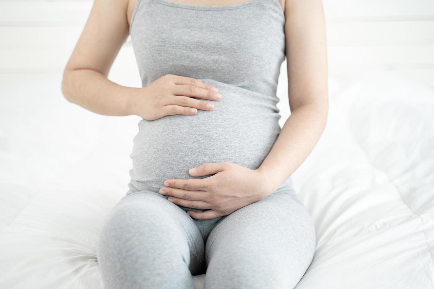 Close up of pregnant woman sitting on the bed and touching her belly at home. Selective focus photo