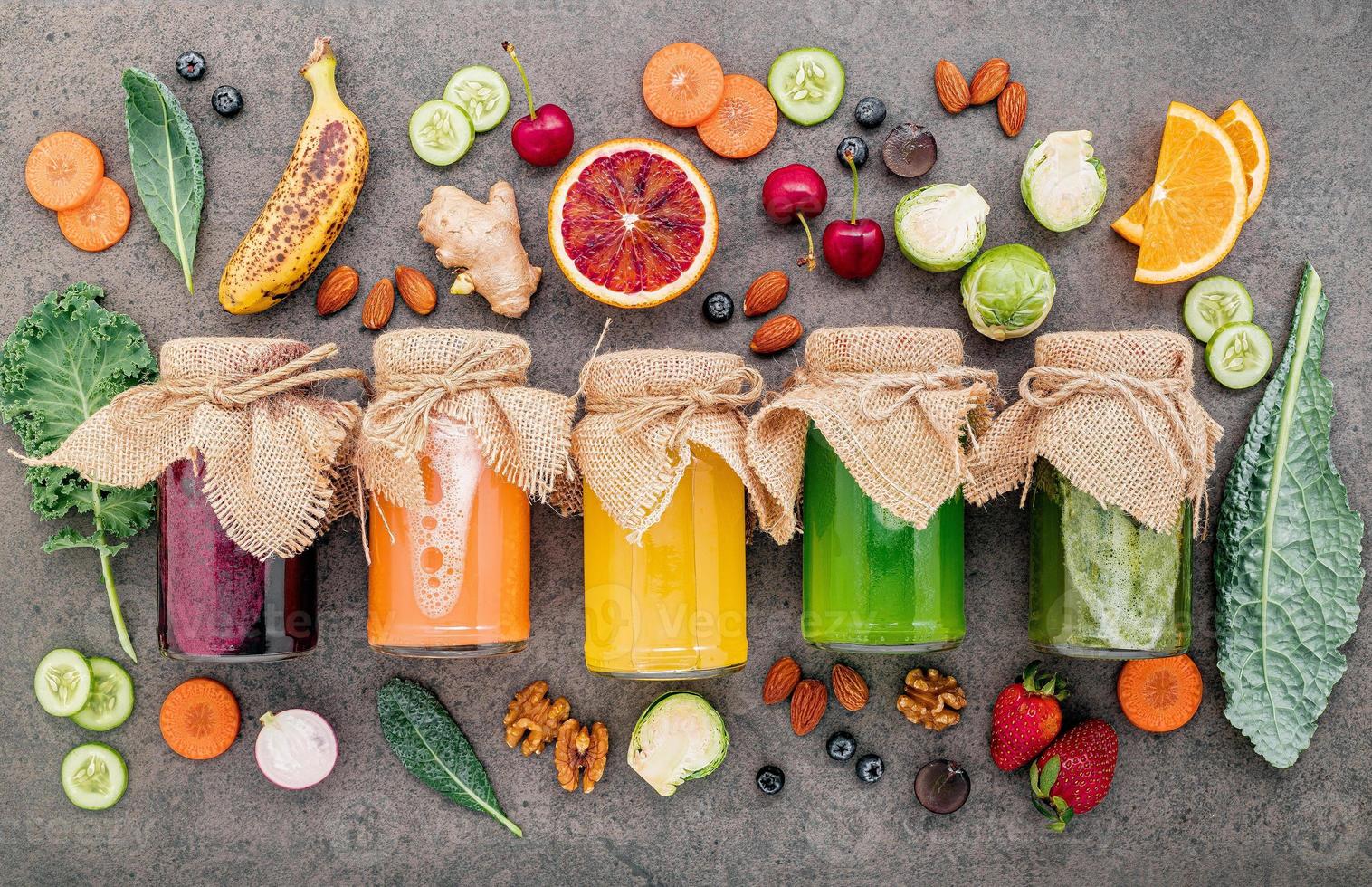 coloridos batidos y jugos saludables en botellas con frutas tropicales frescas y superalimentos sobre fondo de piedra oscura con espacio de copia. foto