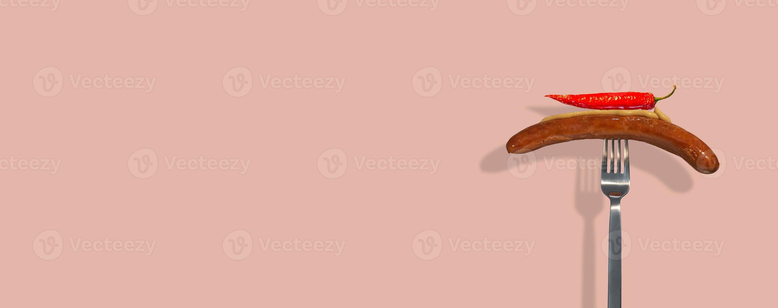 Banner with single grilled sausage with mustard on it and red hot chili pepper put on a metal modern fork at solid pink color background with copy space for text. Concept of street food and cuisine. photo