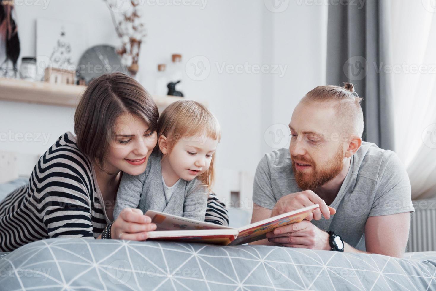 padre, madre e hija leyendo un libro para niños en un sofá en la sala de estar. gran familia feliz lee un libro interesante en un día festivo. los padres aman a sus hijos foto