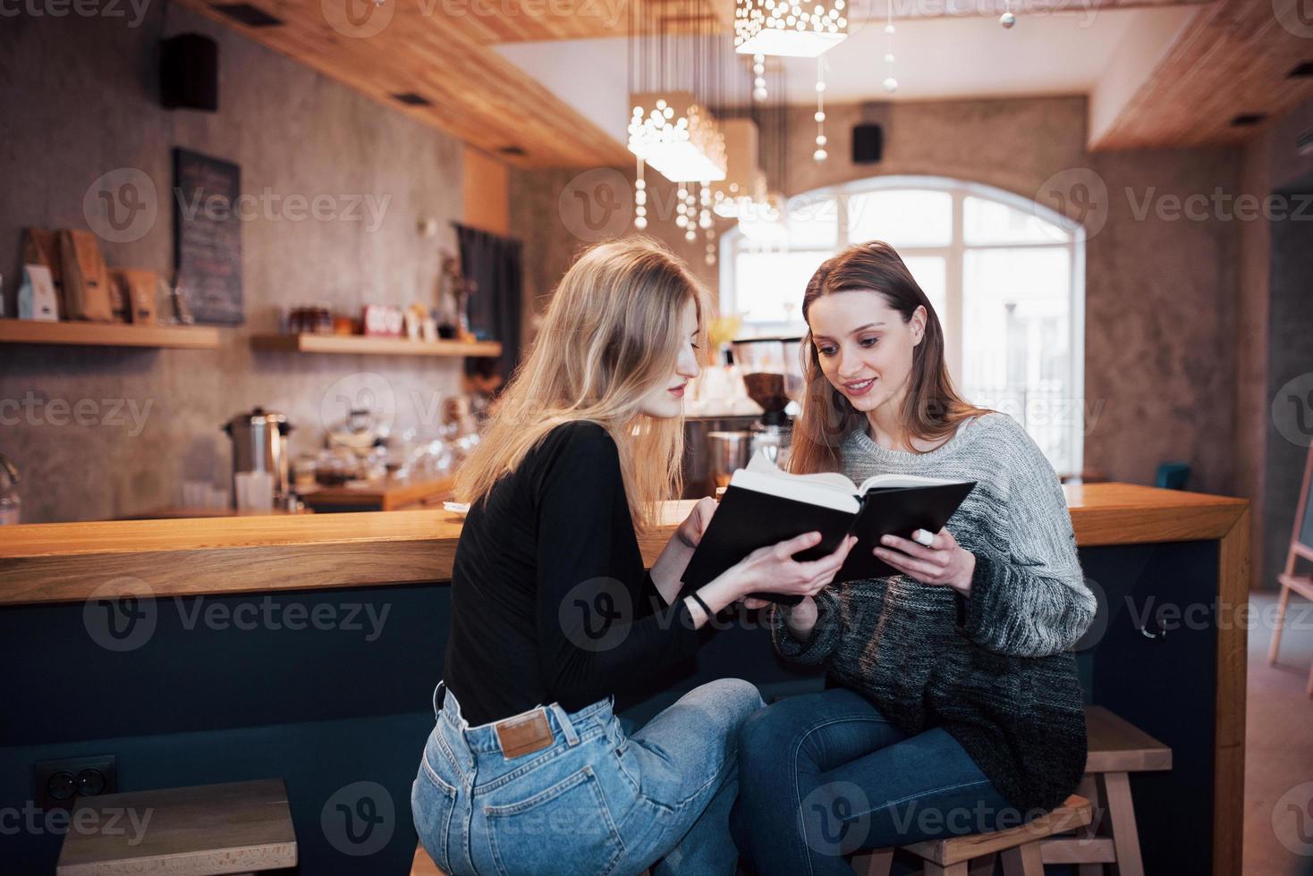 Two Girls absorbed in reading book during the break in cafe. Cute lovely young women are reading book and drinking coffee photo