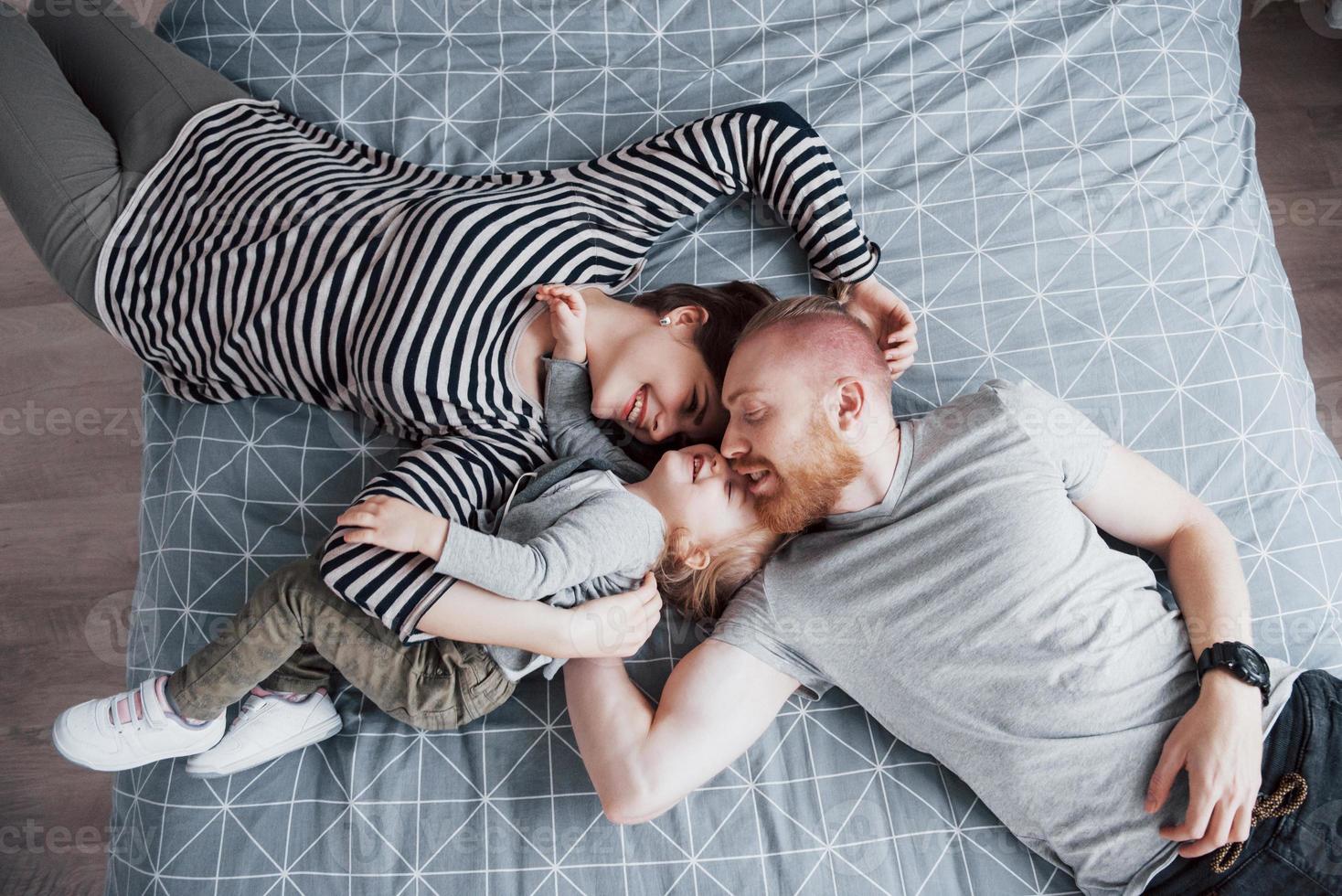 Vista superior de la hermosa joven madre, padre e hija mirando a la cámara y sonriendo mientras está acostado en la cama cabeza a cabeza foto