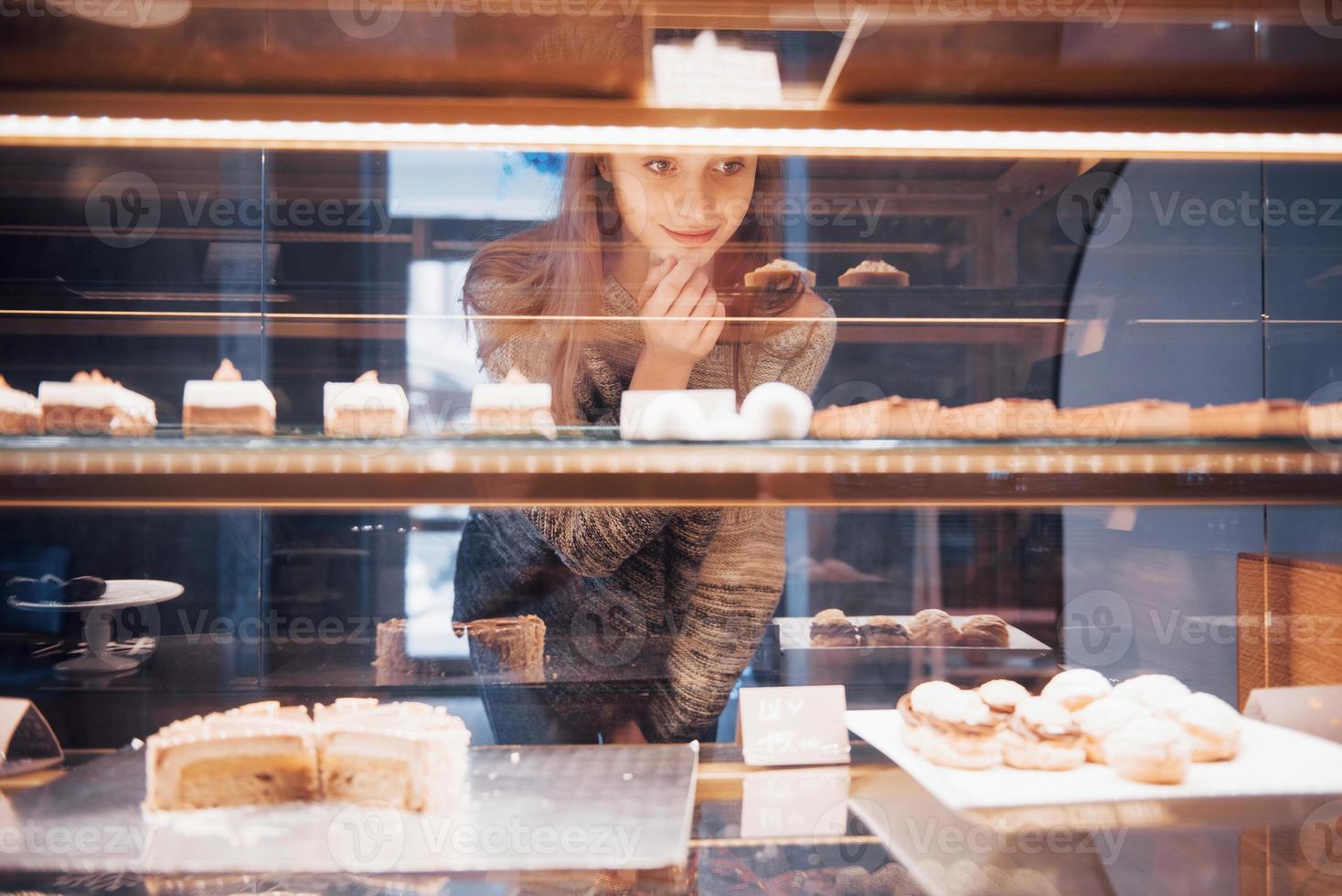 Mujer sonriente a la cámara a través de la vitrina con dulces y tortas en el interior de la cafetería moderna foto