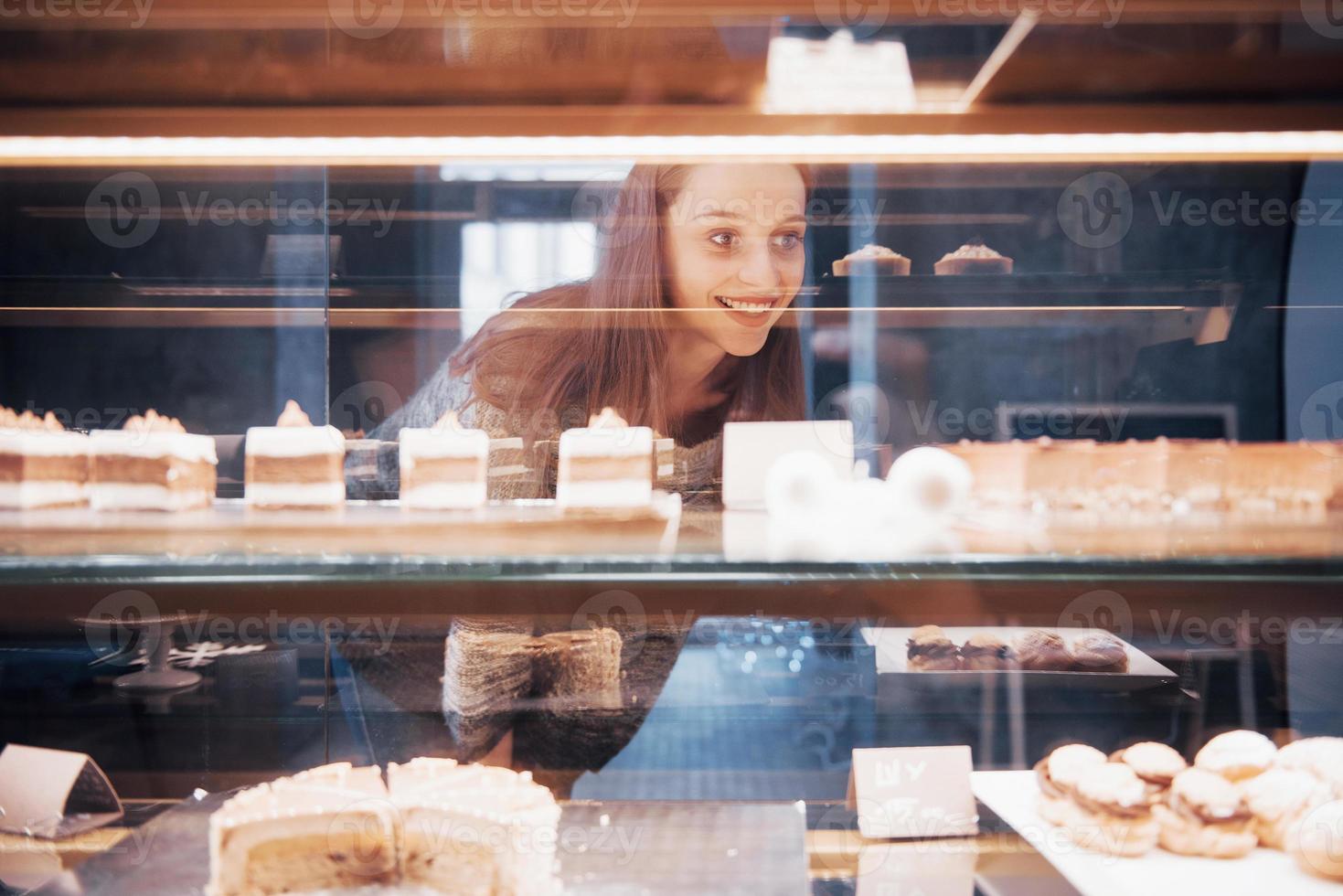 Mujer sonriente a la cámara a través de la vitrina con dulces y tortas en el interior de la cafetería moderna foto