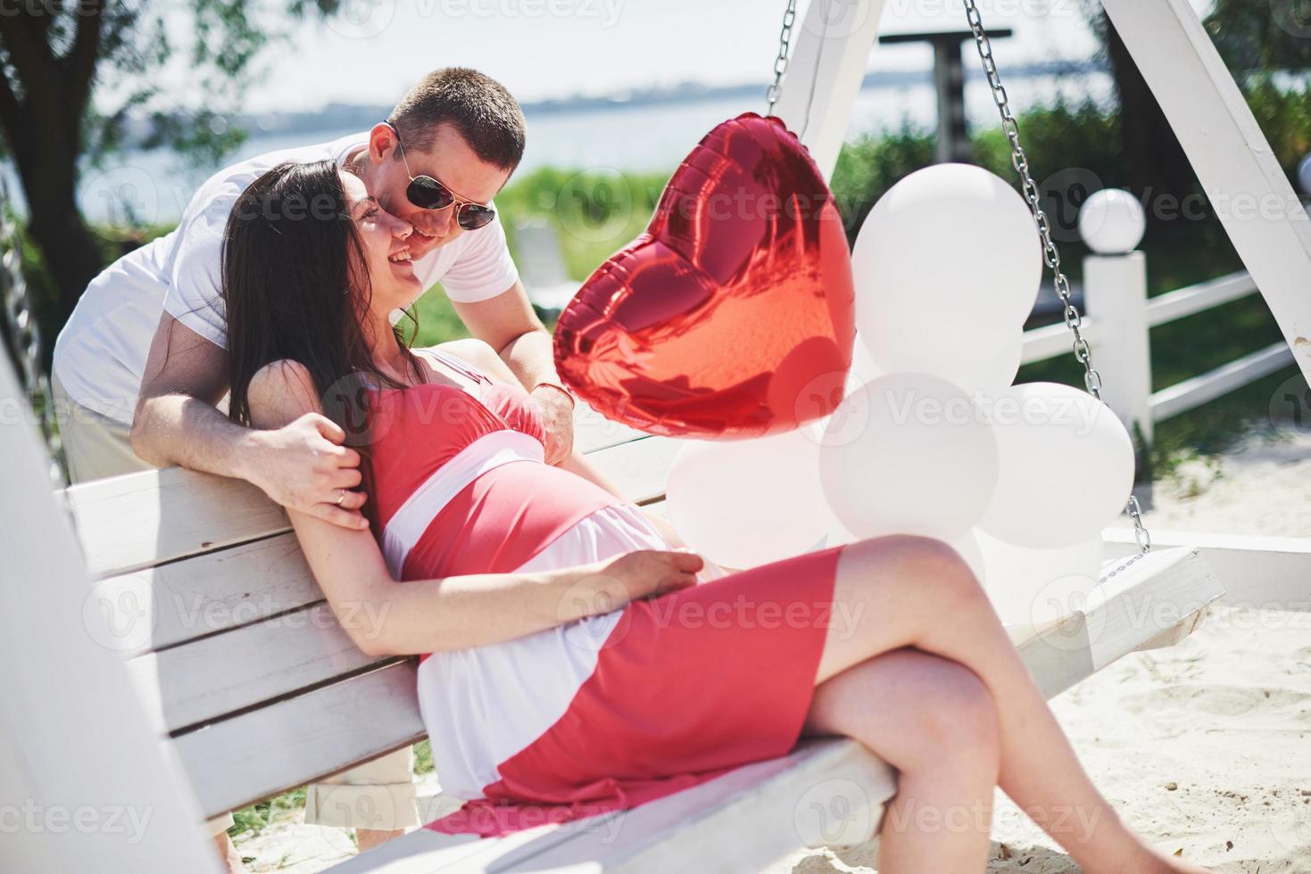 waiting baby. pregnant woman with beloved husband sit on a bench. Husband hand embraces a round belly wife. Parenthood. pregnant woman in a white dress in cherry with a red bow. close-up. nine months photo