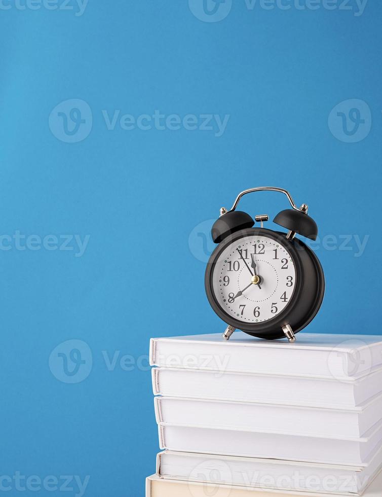 Black alarm clock on stack of books on blue background, mockup design photo