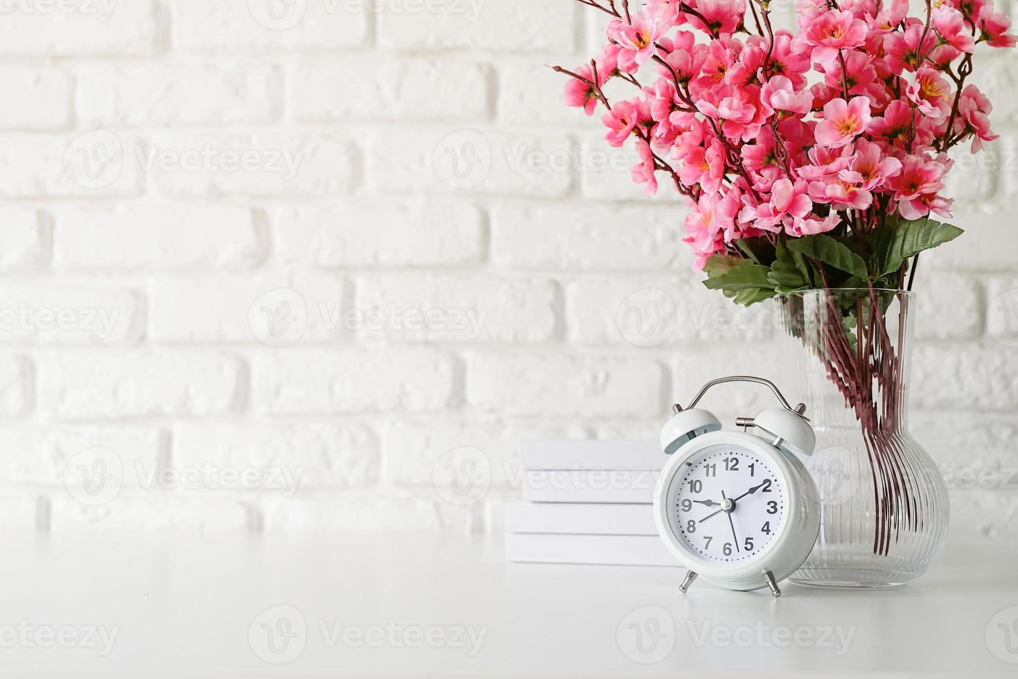 White retro alarm clock on white brick wall with books and flowers with copy space photo