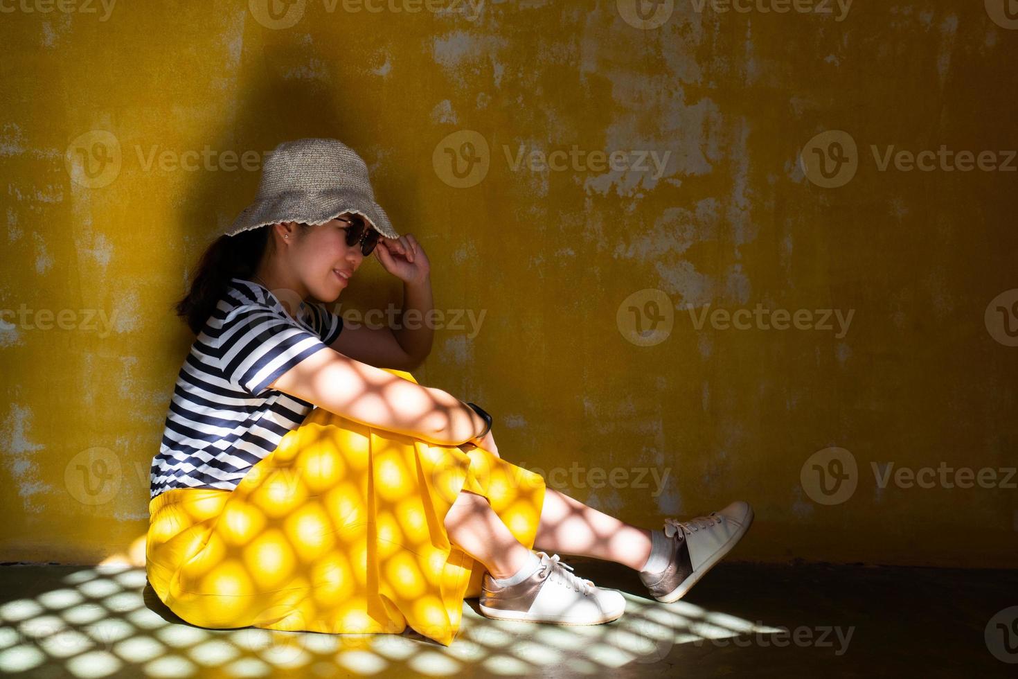 joven y bella mujer asiática con sombrero, gafas de sol y falda amarilla que tiene luz solar en la superficie. Ella se sienta en el suelo con fondo amarillo rayado pelado grunge foto