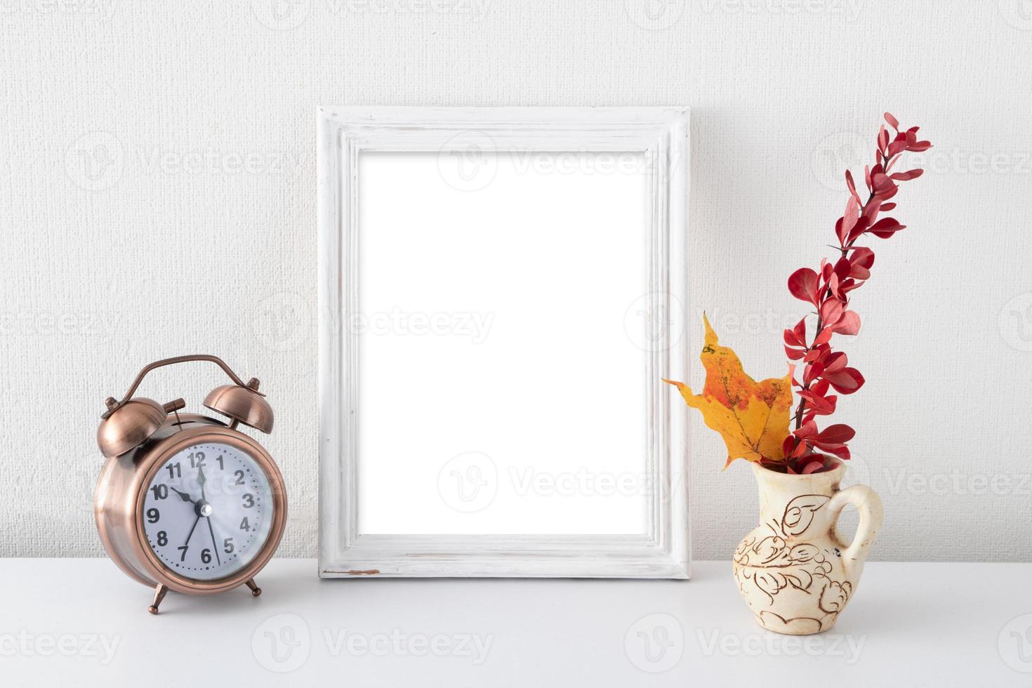 simulacro de marco blanco vacío en el interior blanco cerca de un jarrón con hojas de otoño y reloj despertador foto