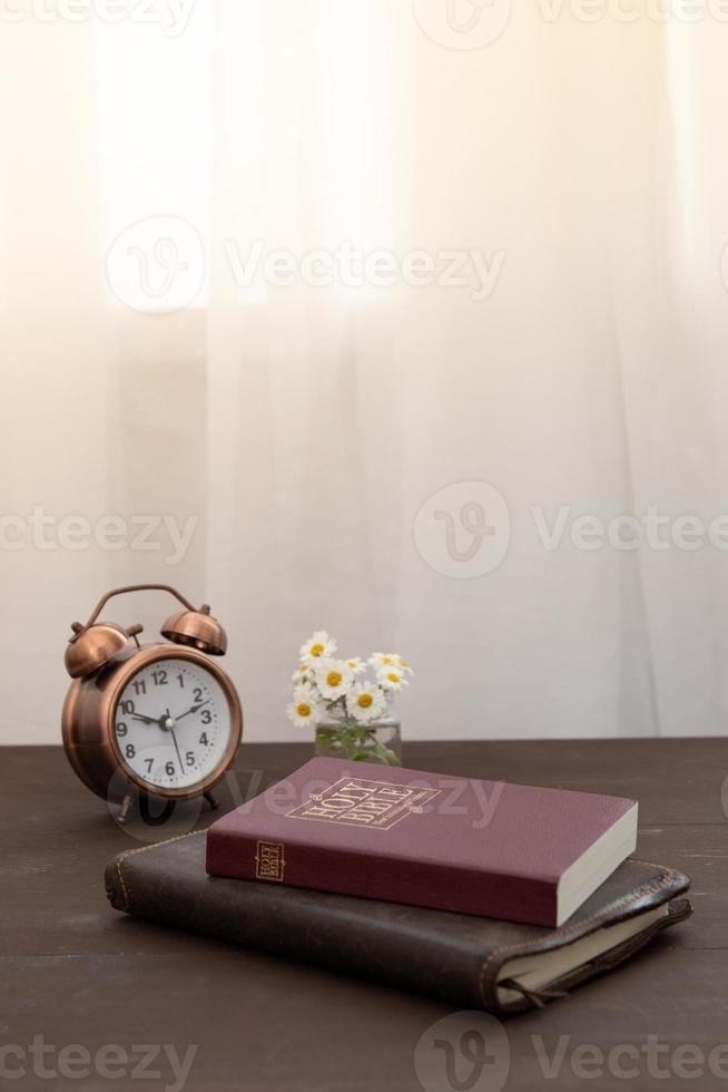 Bible and notebook on the table with an alarm clock by the window. Vertical format photo