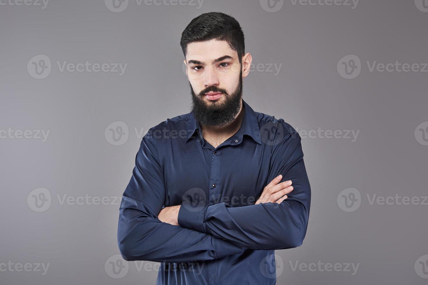 Young handsome man leaning against grey wall with arms crossed. A serious young man with a beard looks at the camera photo