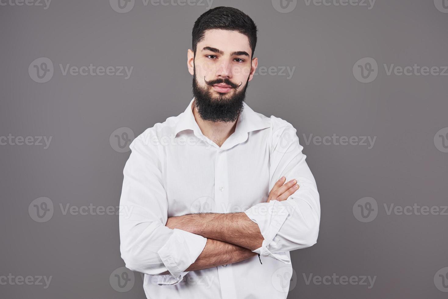Young handsome man leaning against grey wall with arms crossed. A serious young man with a beard looks at the camera photo