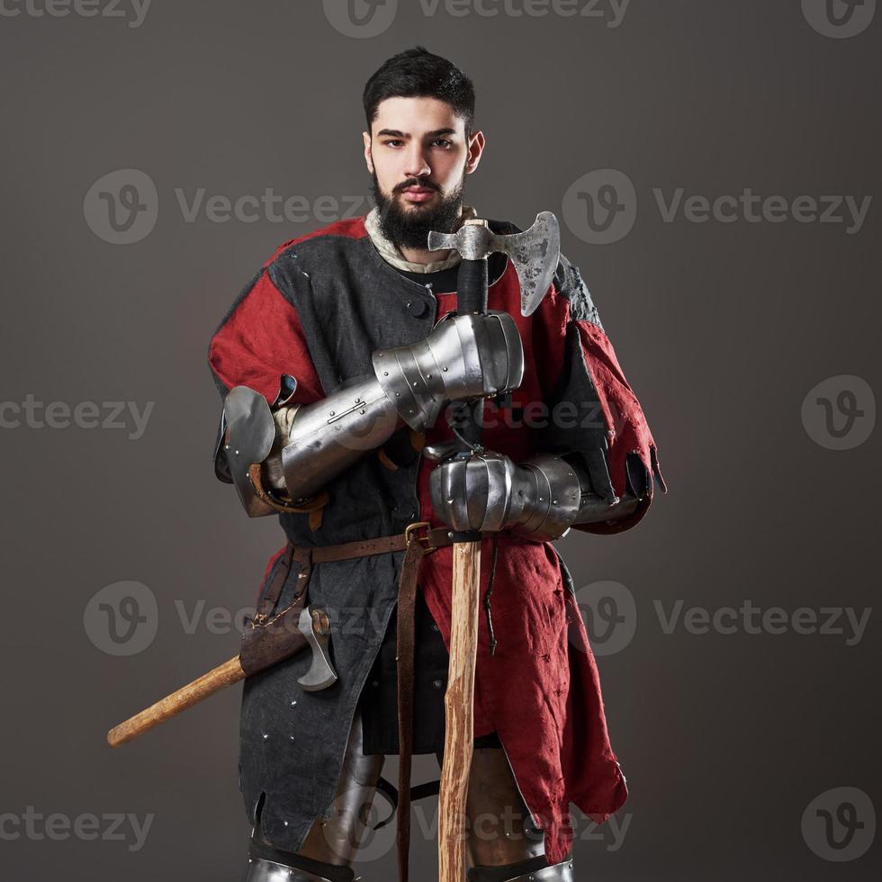 Medieval knight on grey background. Portrait of brutal dirty face warrior with chain mail armour red and black clothes and battle axe photo