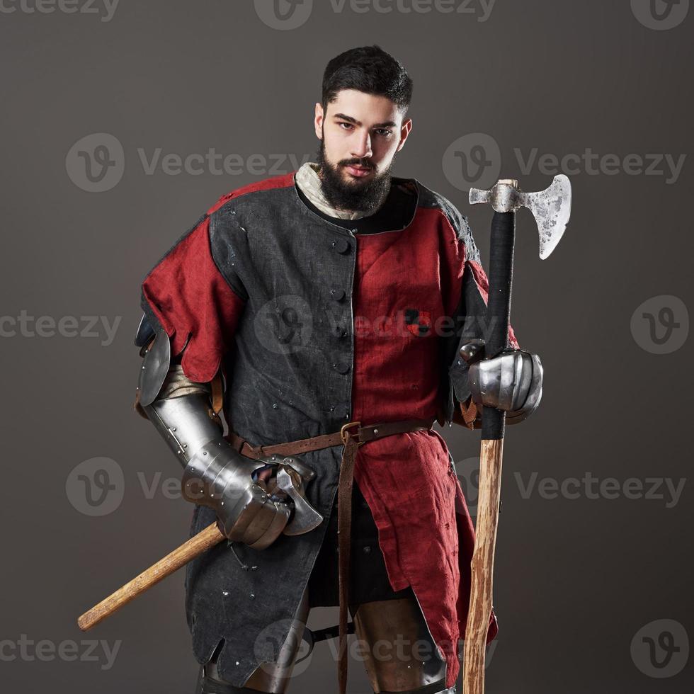 Medieval knight on grey background. Portrait of brutal dirty face warrior with chain mail armour red and black clothes and battle axe photo