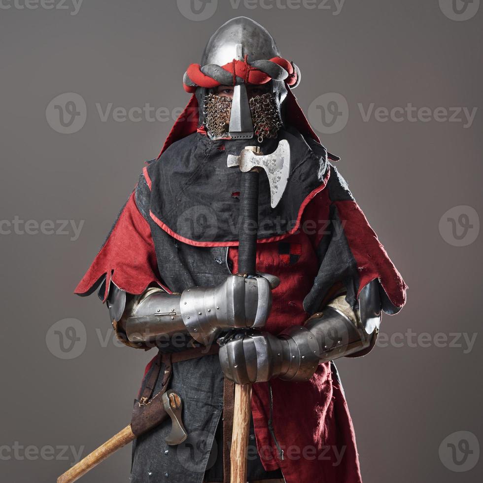 Medieval knight on grey background. Portrait of brutal dirty face warrior with chain mail armour red and black clothes and battle axe photo