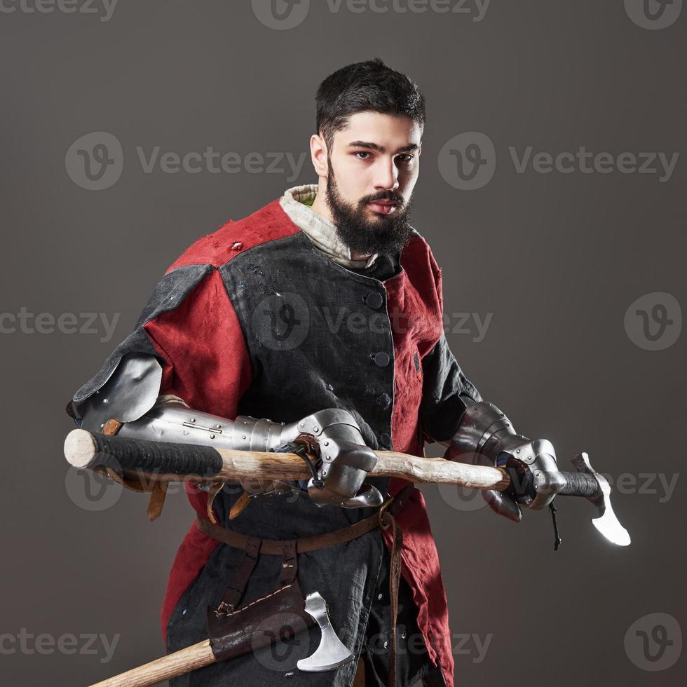 Medieval knight on grey background. Portrait of brutal dirty face warrior with chain mail armour red and black clothes and battle axe photo