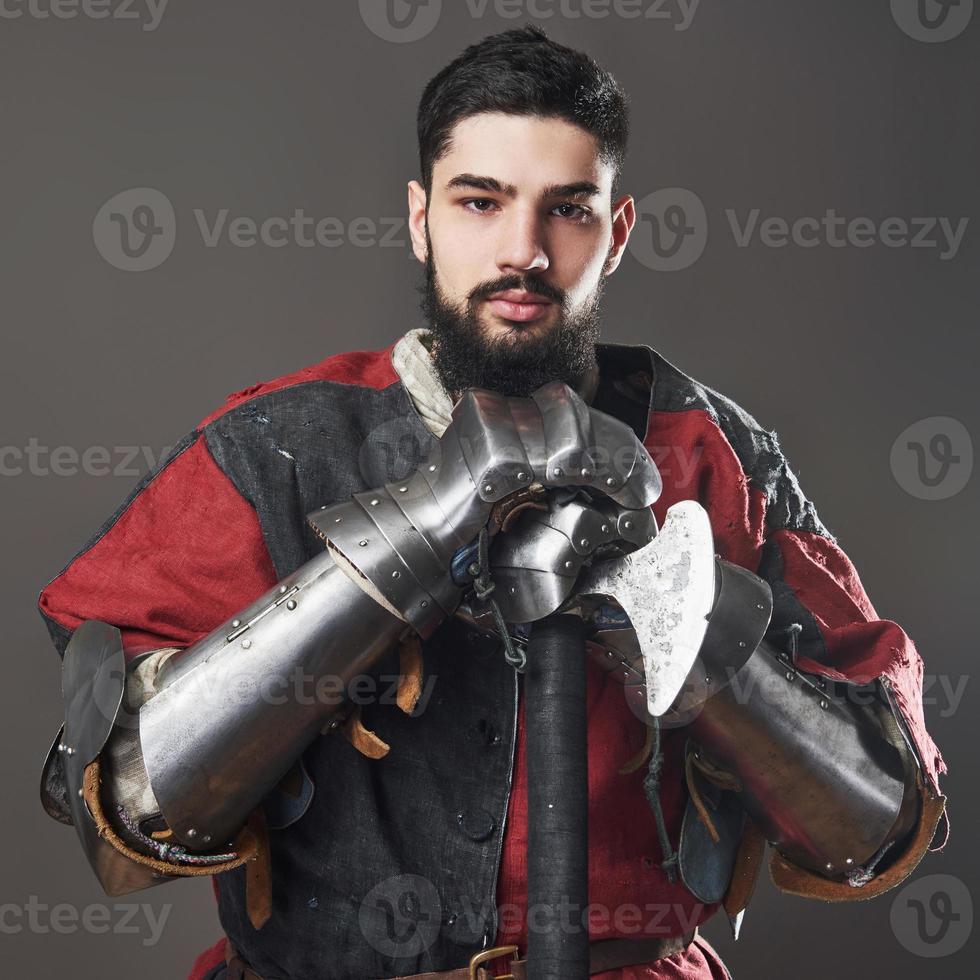 Medieval knight on grey background. Portrait of brutal dirty face warrior with chain mail armour red and black clothes and battle axe photo