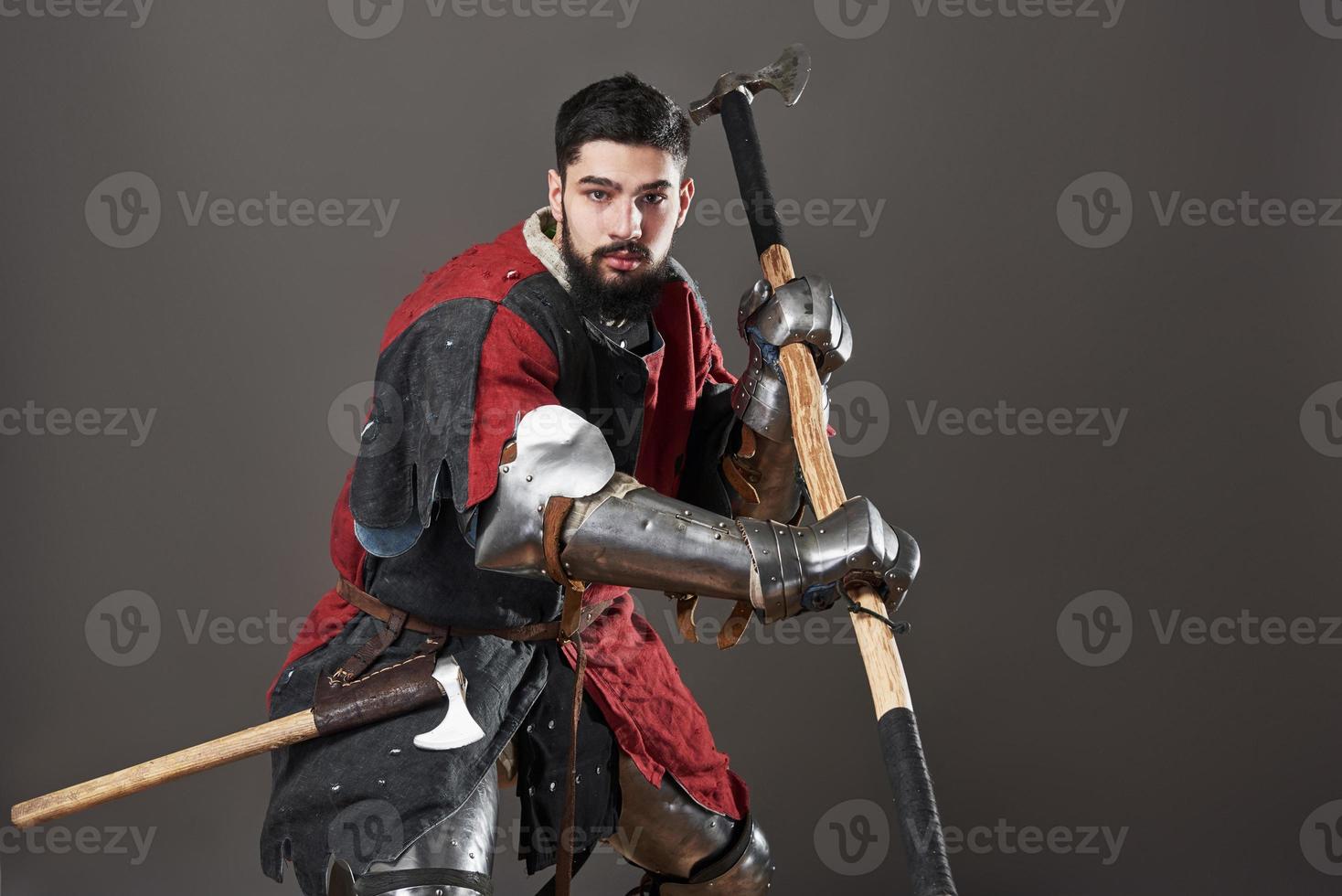 Medieval knight on grey background. Portrait of brutal dirty face warrior with chain mail armour red and black clothes and battle axe photo