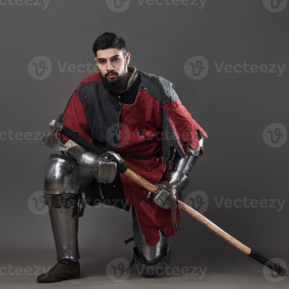 Medieval knight on grey background. Portrait of brutal dirty face warrior with chain mail armour red and black clothes and battle axe photo