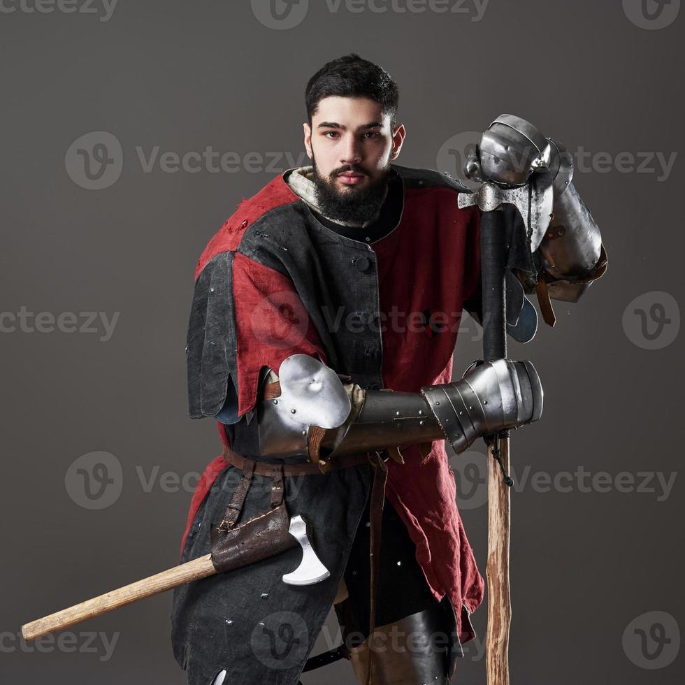 Medieval knight on grey background. Portrait of brutal dirty face warrior with chain mail armour red and black clothes and battle axe photo