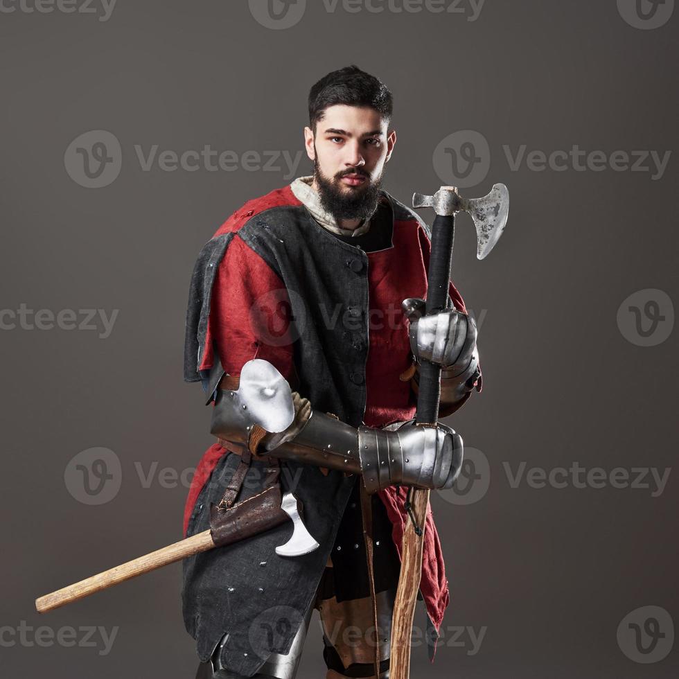 Medieval knight on grey background. Portrait of brutal dirty face warrior with chain mail armour red and black clothes and battle axe photo