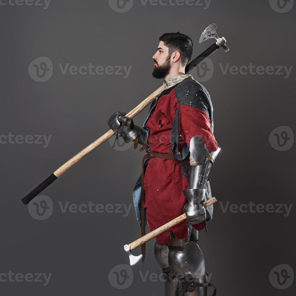 Medieval knight on grey background. Portrait of brutal dirty face warrior with chain mail armour red and black clothes and battle axe photo