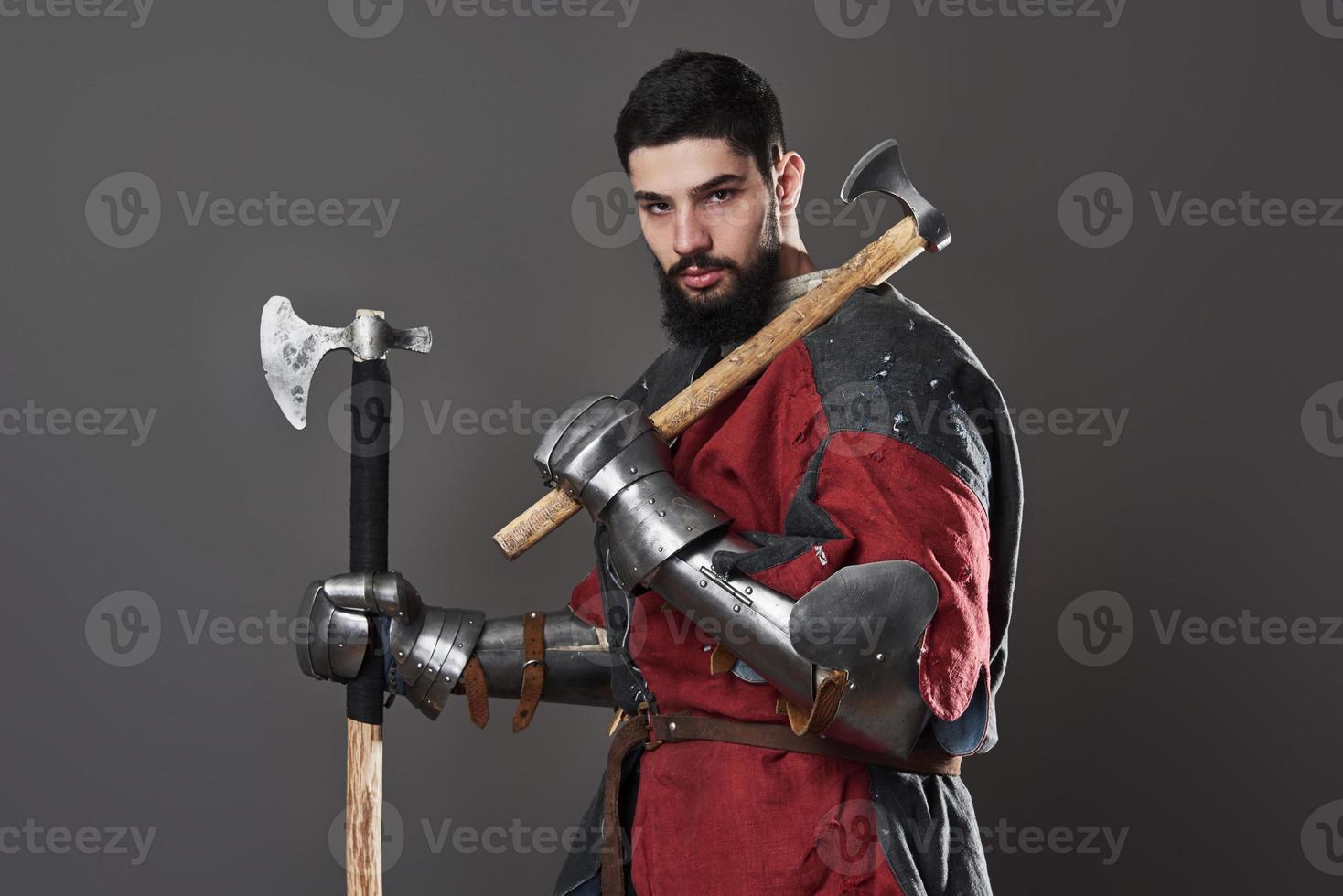 Medieval knight on grey background. Portrait of brutal dirty face warrior with chain mail armour red and black clothes and battle axe photo