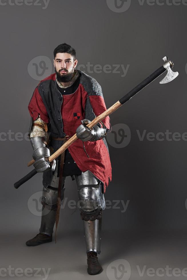 Medieval knight on grey background. Portrait of brutal dirty face warrior with chain mail armour red and black clothes and battle axe photo