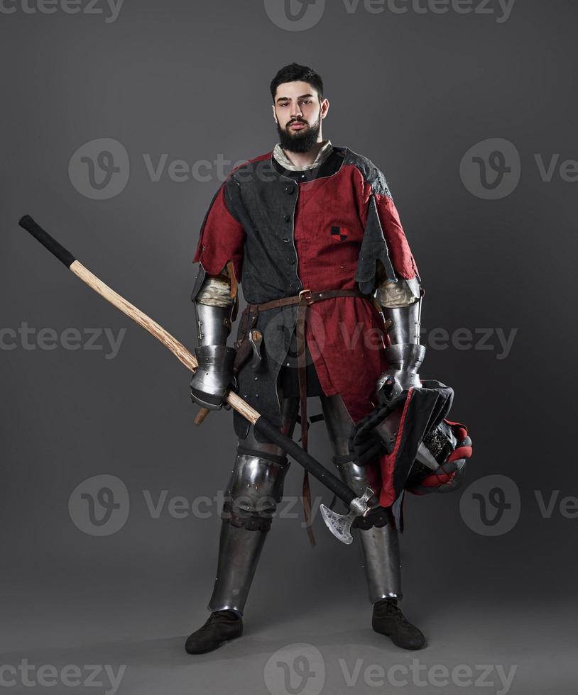 Medieval knight on grey background. Portrait of brutal dirty face warrior with chain mail armour red and black clothes and battle axe photo