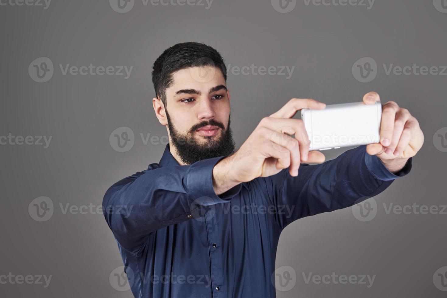 Young bearded hipster business man talking selfie photo with smart phone smiling and looking at phone against gray studio background