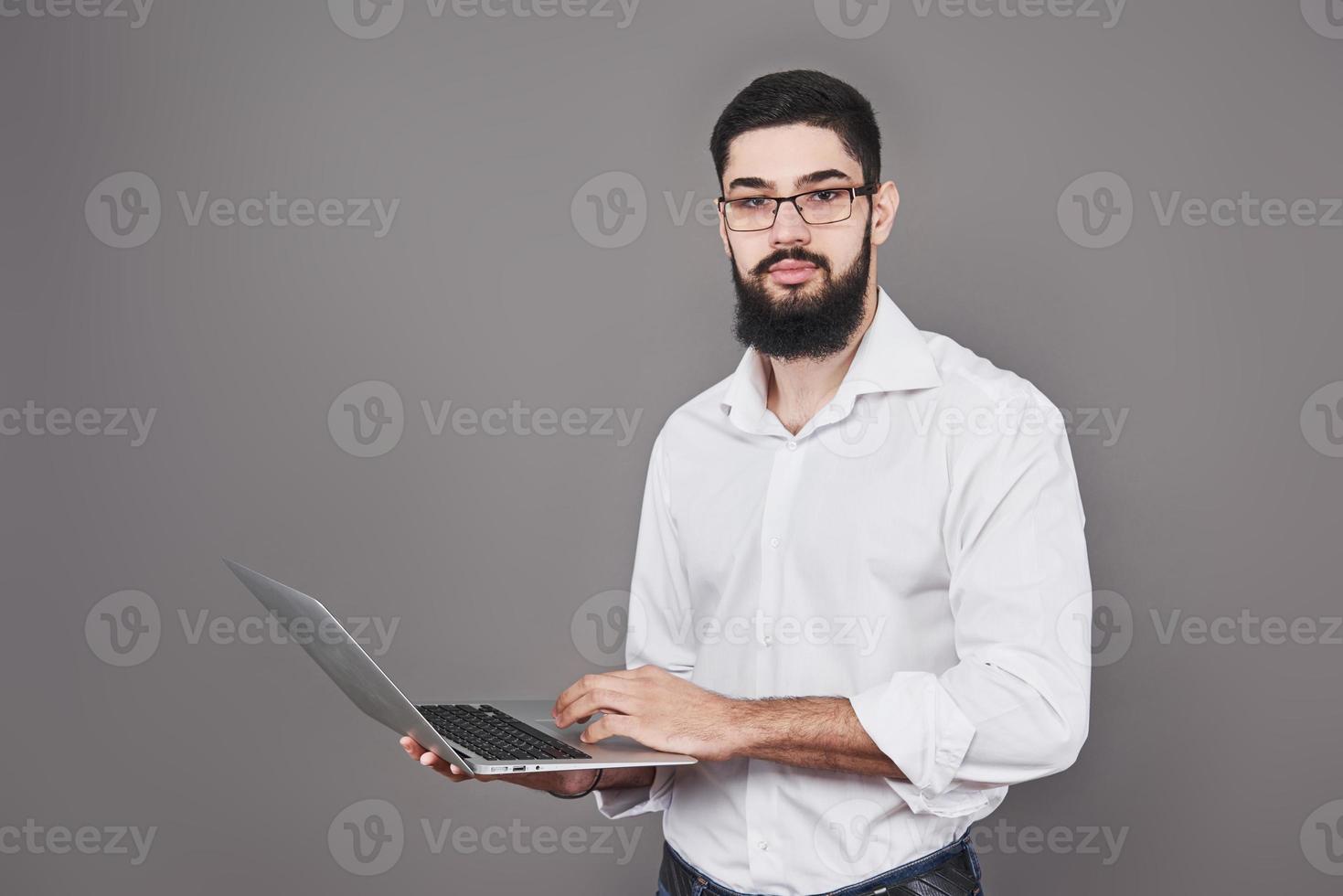 apuesto hombre de negocios con gafas y traje sosteniendo una computadora portátil en las manos y escribiendo algo. vista lateral. fondo gris aislado foto