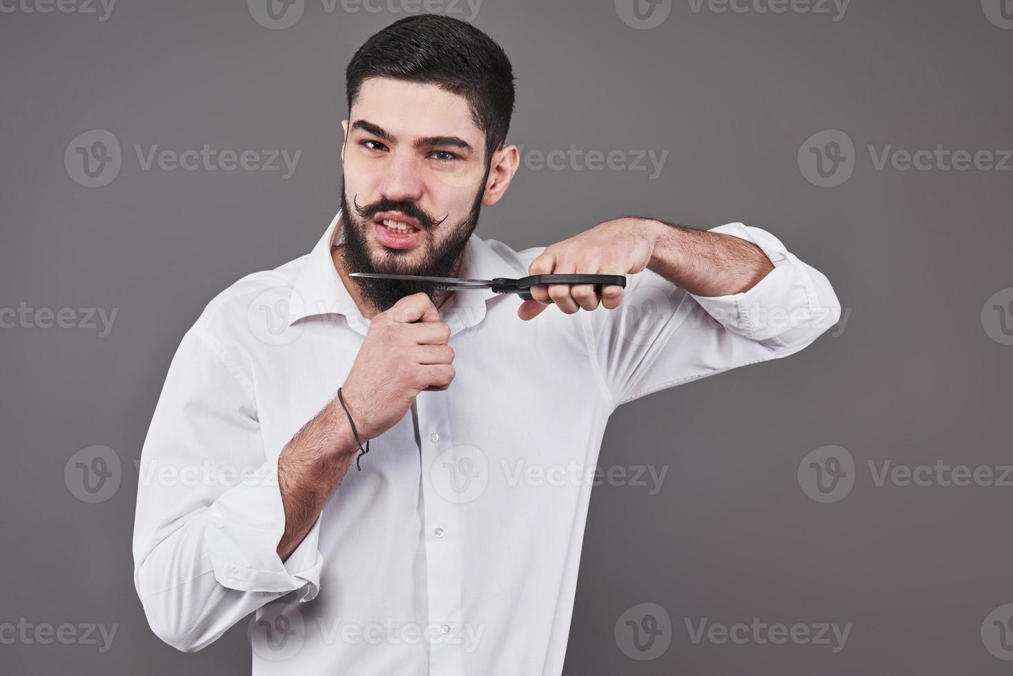 No more beard. Portrait of handsome young man cutting his beard with scissors and looking at camera while standing against grey background. New trend photo