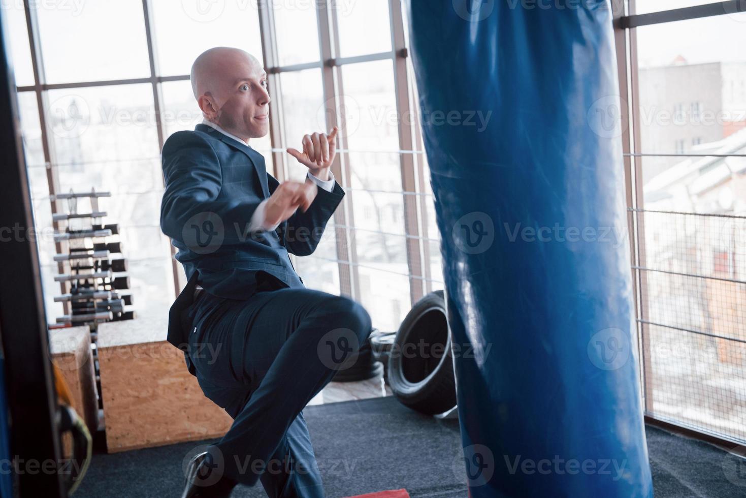 un hombre de negocios calvo enojado golpea una pera de boxeo en el gimnasio. concepto de manejo de la ira foto