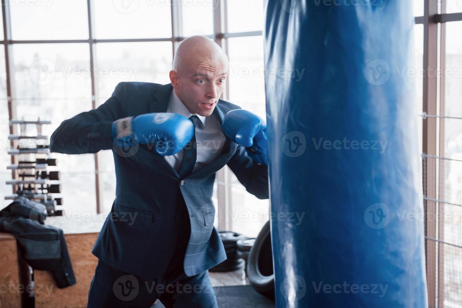 un hombre de negocios calvo enojado golpea una pera de boxeo en el gimnasio. concepto de manejo de la ira foto