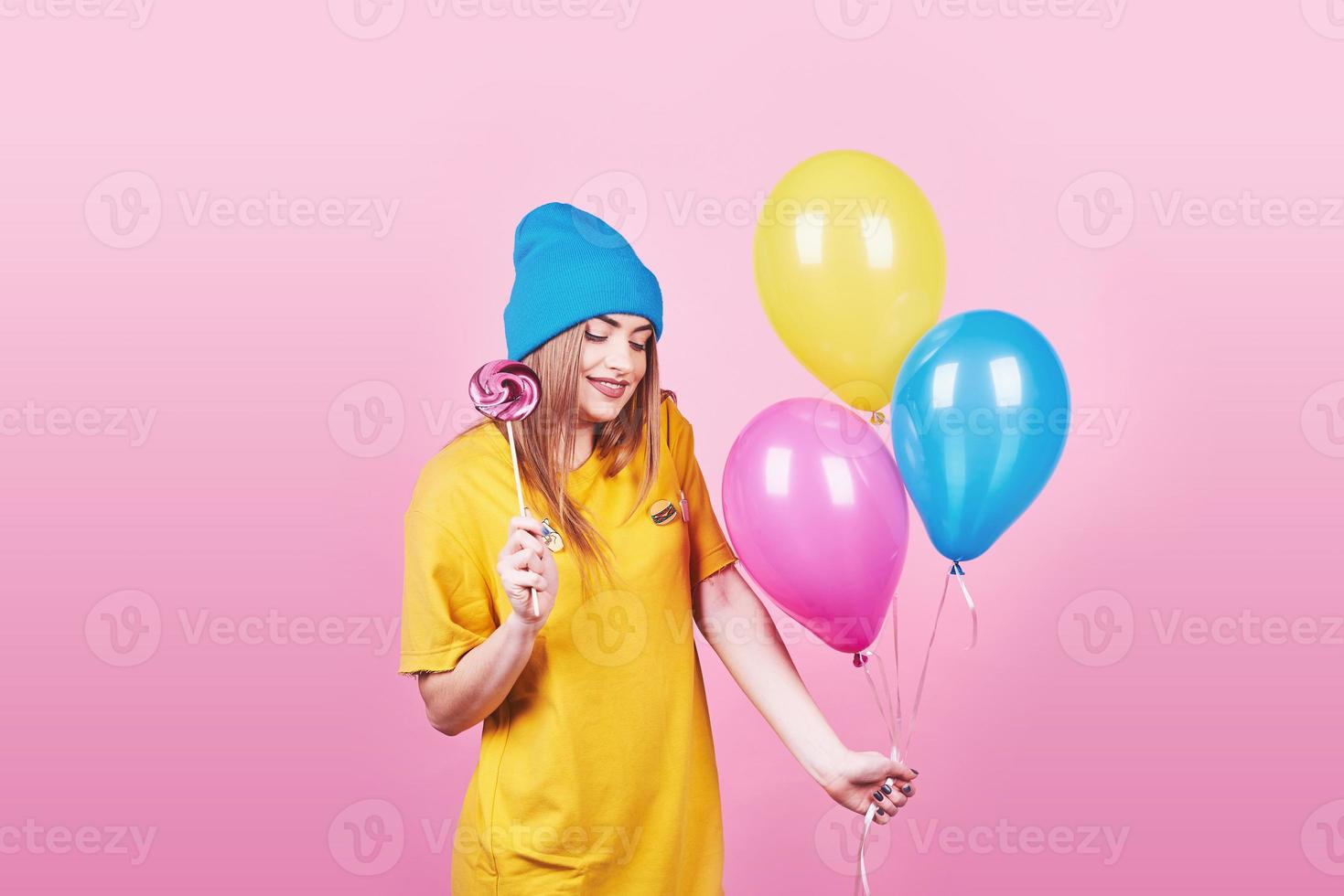 Cute funny girl in blue cap portrait holds an air colorful balloons and lollipop smiling on pink background. Beautiful multicultural Caucasian girl smiling happy photo
