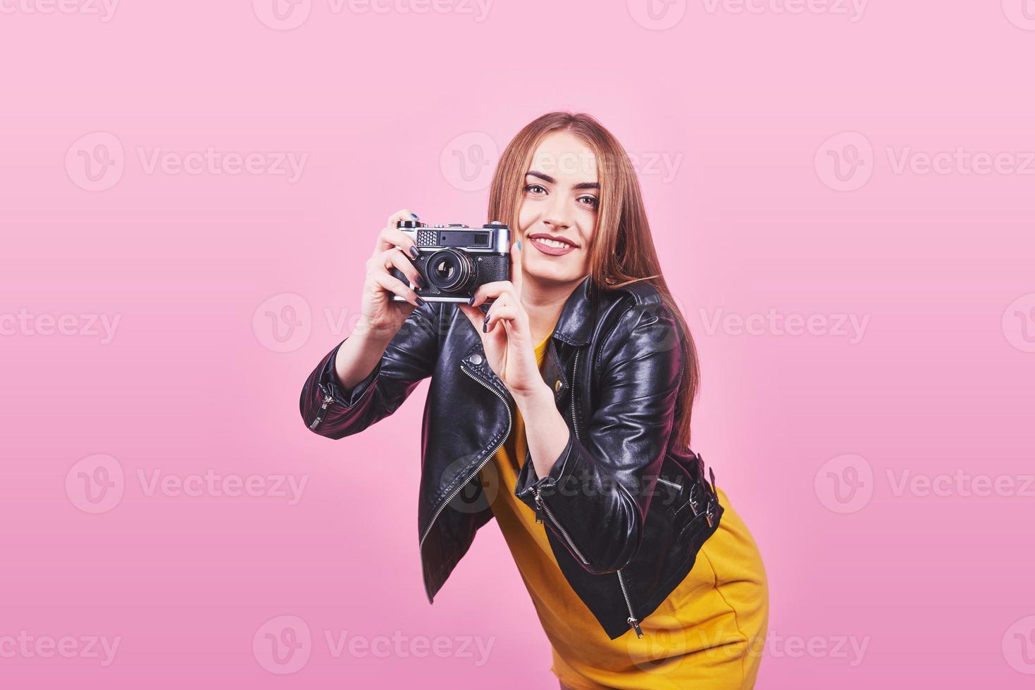 Fashion look, pretty cool young woman model with retro film camera wearing a black jacket, against pink background with copy space photo