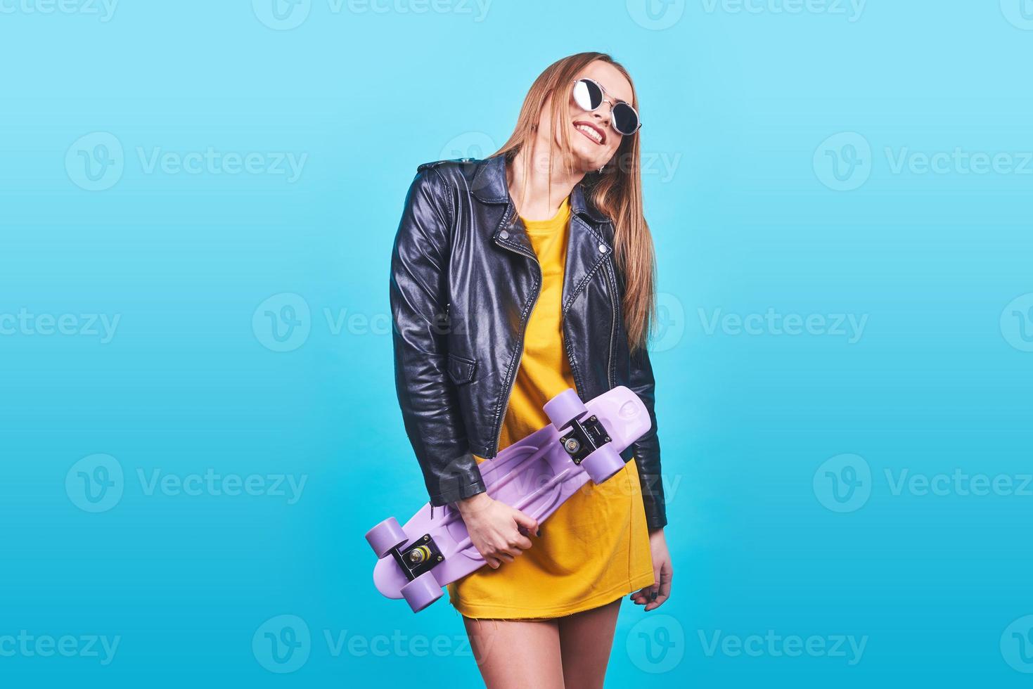 Attractive tanned girl with smile in face wears black leather jacket posing with pleasure. Indoor photo of lovely woman in sunglasses standing with skateboard on blue background