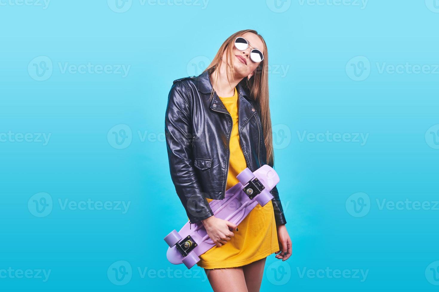 atractiva chica bronceada con sonrisa en la cara viste chaqueta de cuero negro posando con placer. Fotografía interior de una mujer encantadora en gafas de sol de pie con patineta sobre fondo azul. foto