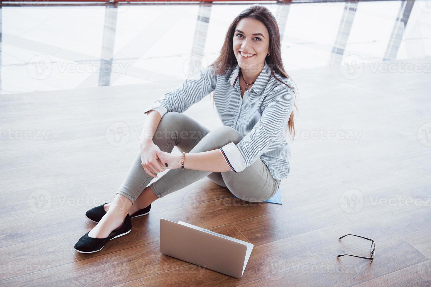 Side view of attractive girl using a laptop in public wifi area and smiling while sitting on the floor photo