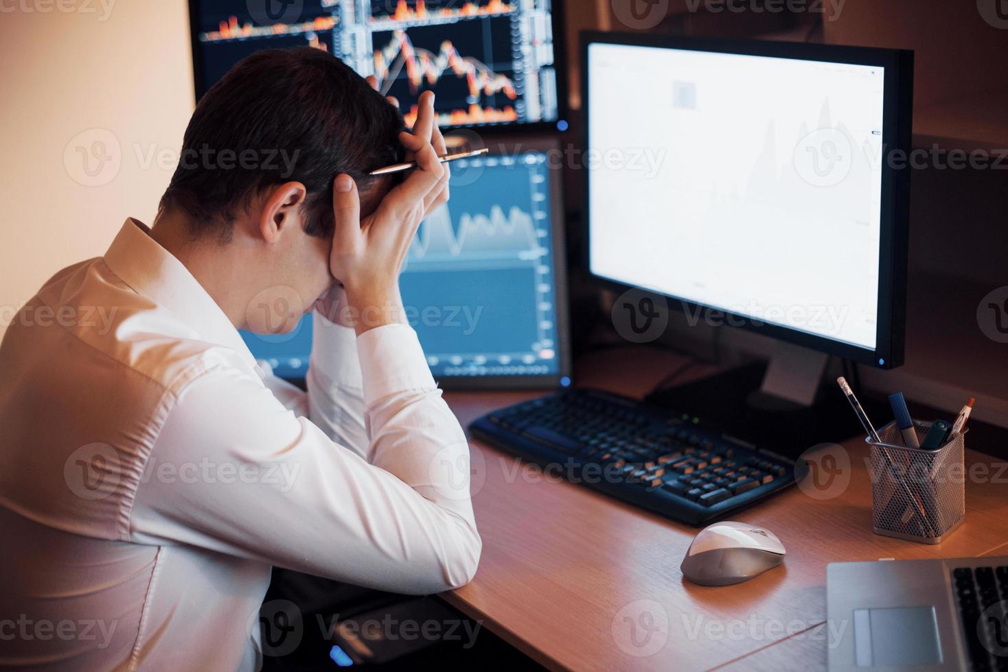 Stressful day at the office. Young businessman holding hands on his face while sitting at the desk in creative office. Stock Exchange Trading Forex Finance Graphic Concept photo