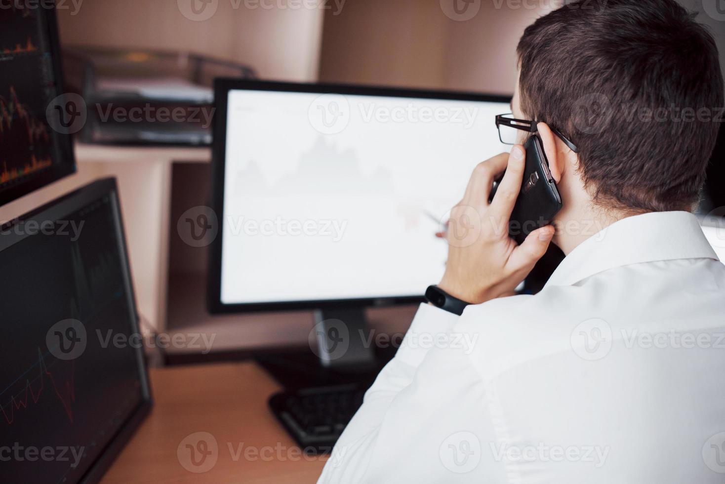 Over the shoulder view of and stock broker trading online while accepting orders by phone. Multiple computer screens ful of charts and data analyses in background photo