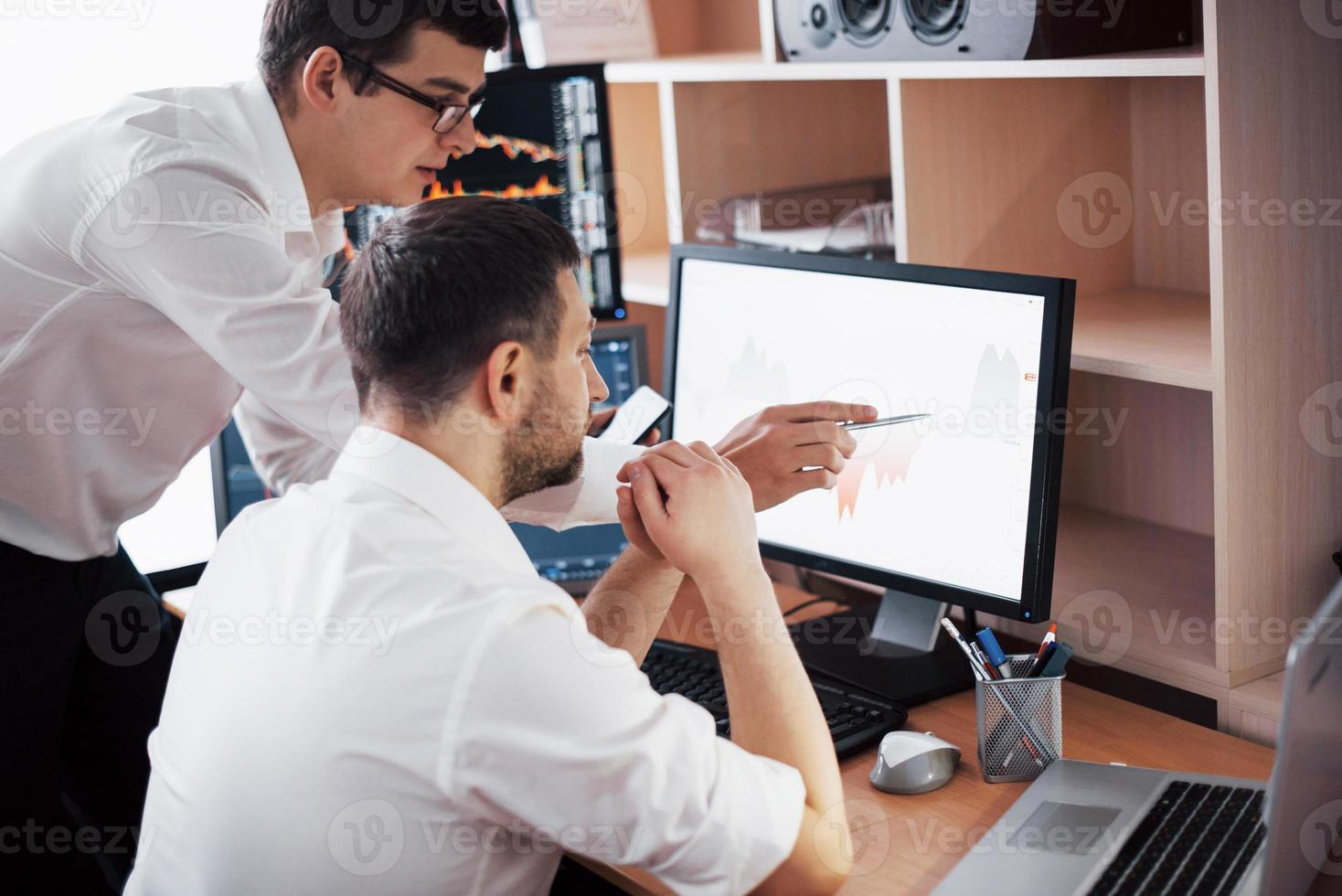 Businessmen trading stocks online. Stock brokers looking at graphs, indexes and numbers on multiple computer screens. Colleagues in discussion in traders office. Business success concept photo