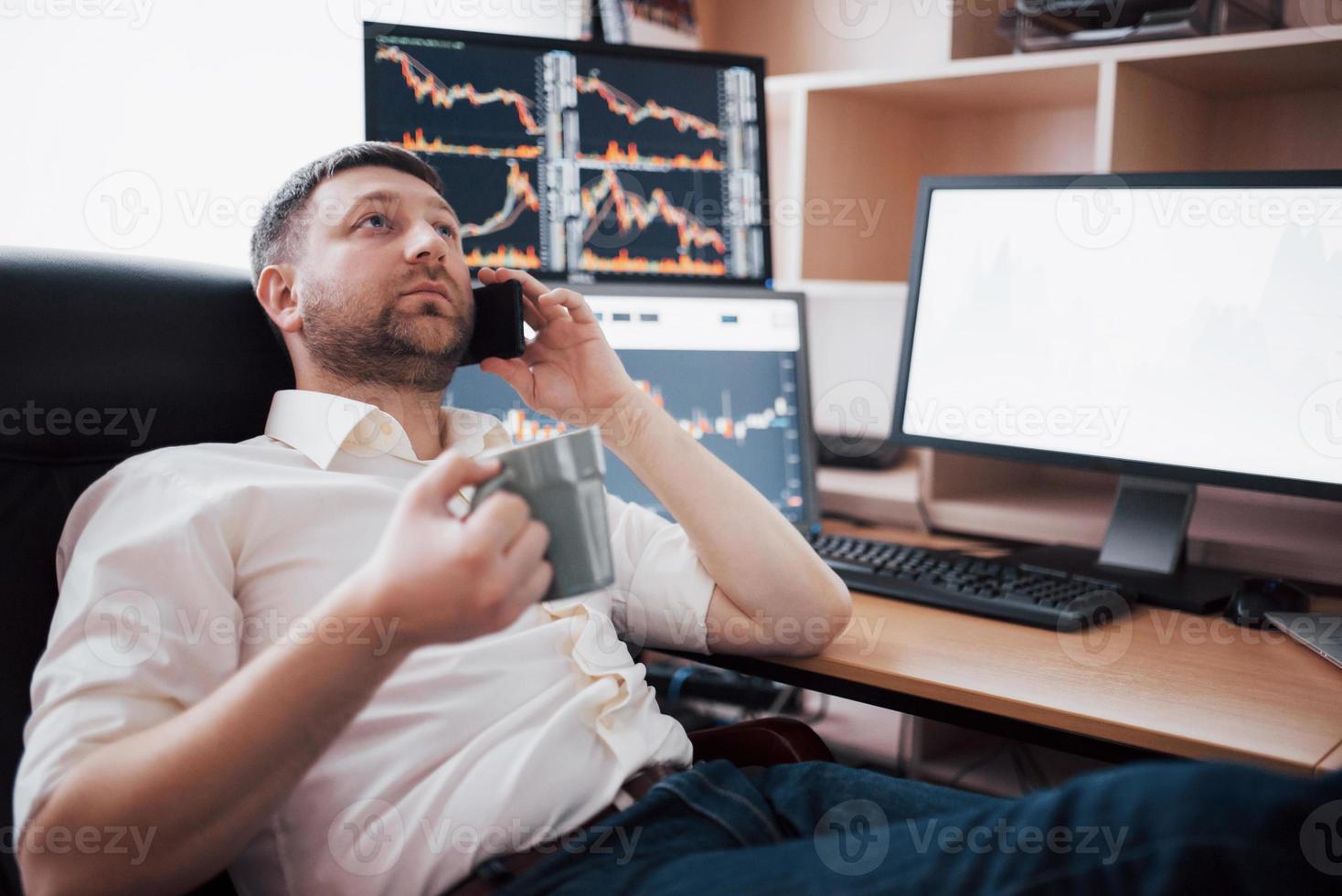 Over the shoulder view of and stock broker trading online while accepting orders by phone. Multiple computer screens ful of charts and data analyses in background photo