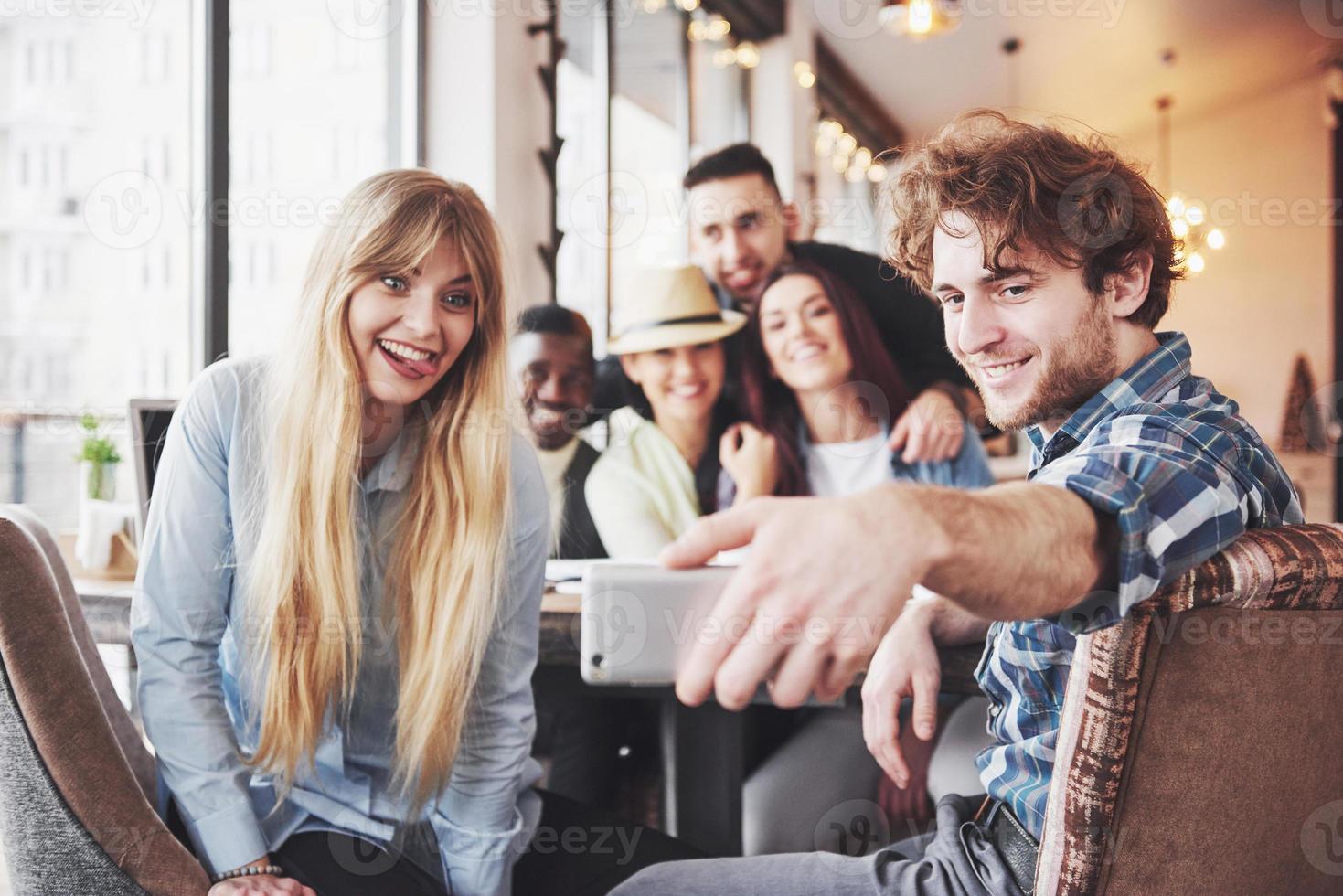 amigos divirtiéndose en el restaurante dos niños y cuatro niñas bebiendo haciendo selfie, haciendo el signo de la paz y riendo. en primer plano mujer sosteniendo teléfono inteligente. todos usan ropa casual foto