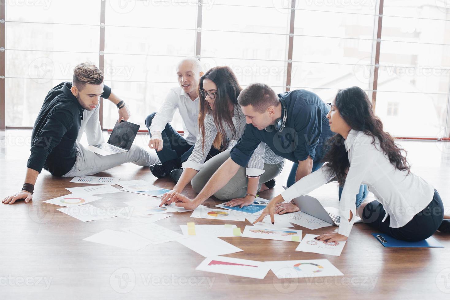 Planning strategy together. Business team looking at papers on floor with manager pointing to one idea. Cooperation corporate achievement. Planning design draw. Teamwork concept photo