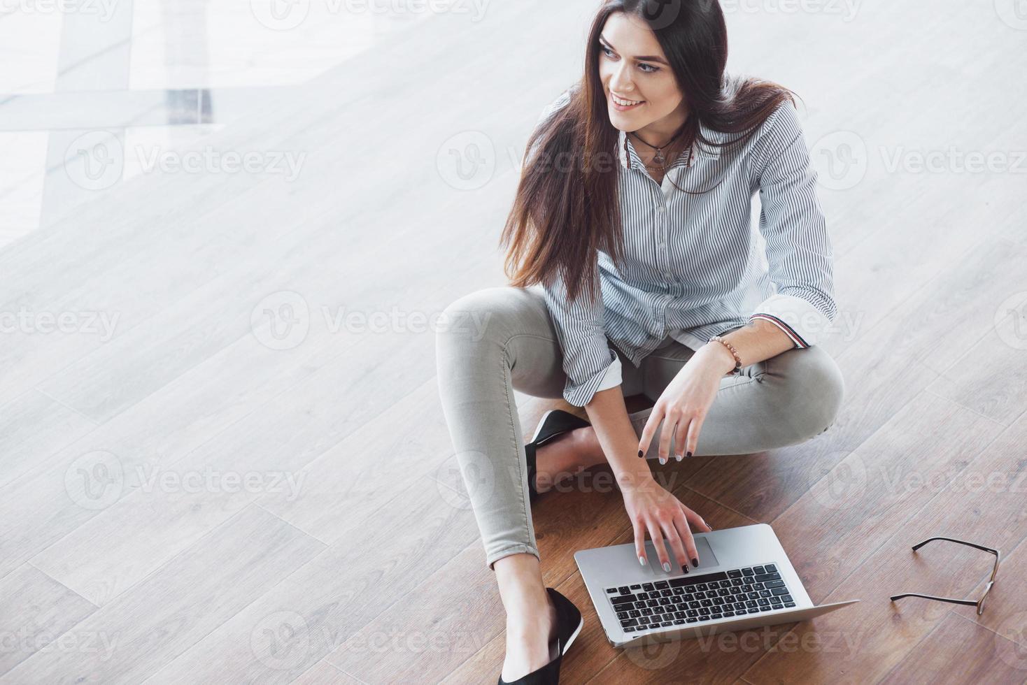 Vista lateral de una chica atractiva usando una computadora portátil en el área wifi pública y sonriendo mientras está sentada en el piso foto