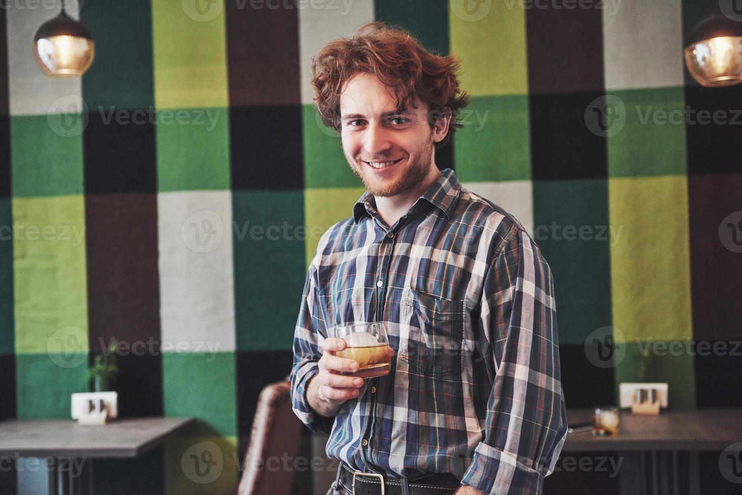 Chico divertido sosteniendo un vaso de whisky y posando contra el papel pintado vintage foto