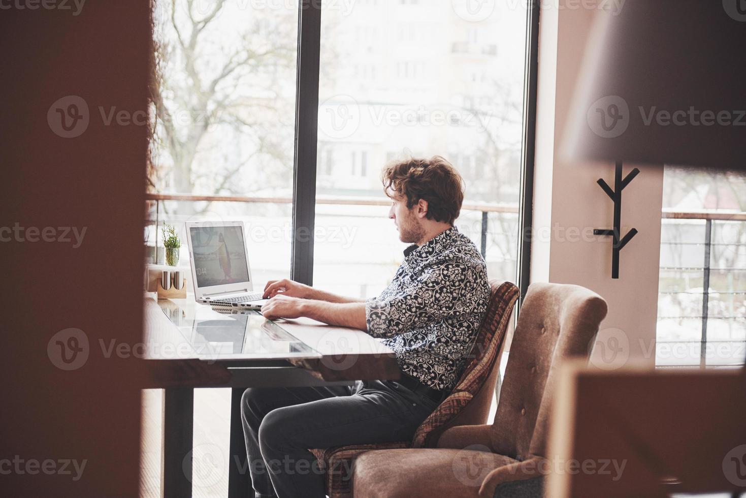 Young handsome man sitting in office with cup of coffee and working on project connected with modern cyber technologies. Businessman with notebook trying to keep deadline in digital marketing sphere photo