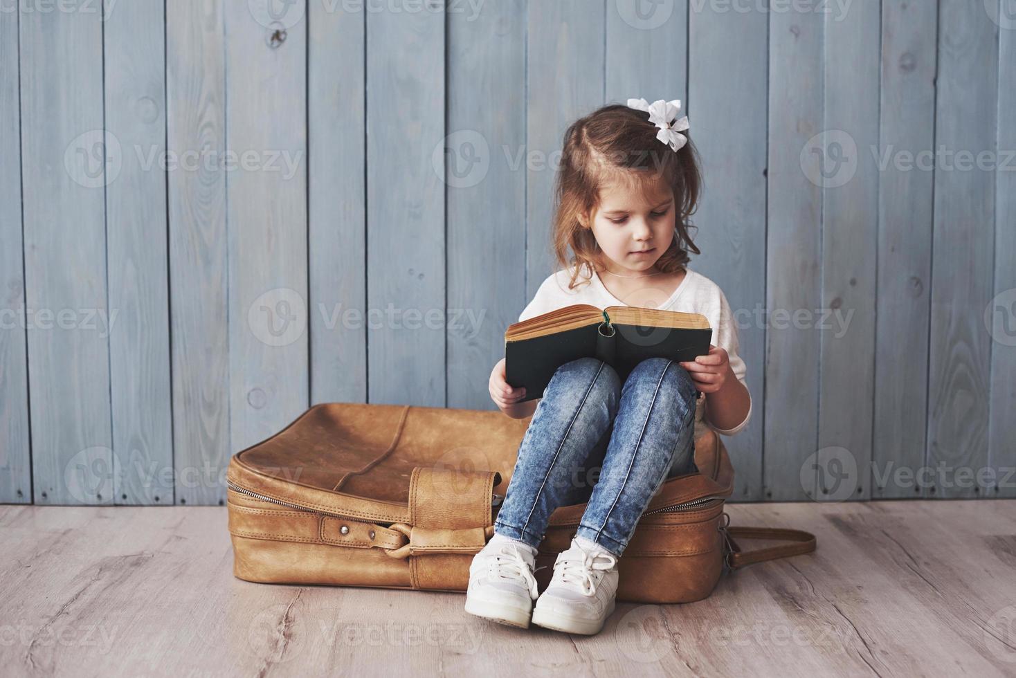 listo para grandes viajes. niña feliz leyendo un libro interesante con un maletín grande y sonriendo. concepto de viaje, libertad e imaginación. foto